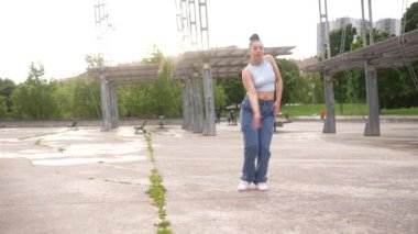 Bir kadın beton zeminde dans ediyor. O, kot pantolon ve mavi bir gömlek giyiyor. Manzara enerjik ve eğlenceli.