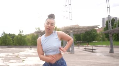 Bir kadın beton zeminde dans ediyor. O, kot pantolon ve mavi bir gömlek giyiyor. Manzara enerjik ve eğlenceli.