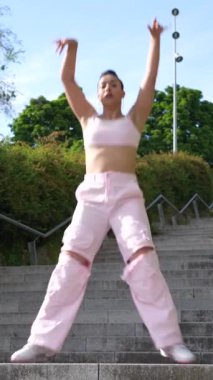 Pembe pantolonlu bir kadın merdivenlerde dans ediyor. Pembe bir bluz giyiyor ve ellerini havaya kaldırıyor. Özgürlük ve neşe kavramı, bir kadının dans ederek kendini ifade etmesi gibi.