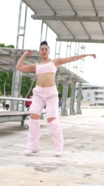 Mavi kolsuz bluzlu ve kot pantolonlu bir kadın bir bankın önünde duruyor. Kameraya bakıyor ve dalgın bir ruh hali içinde.