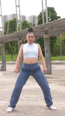 Mavi kolsuz bluzlu ve kot pantolonlu bir kadın binanın önünde dans ediyor. Sahne neşeli ve eğlenceli, çünkü kadın dans ederken eğleniyor.