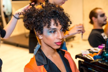 Siyah turuncu elbiseli bir kadın saçını yaptırıyor. Kırmızı bir dudağı var ve mavi göz farı sürüyor. Cadılar Bayramı partisi hazırlıkları için.