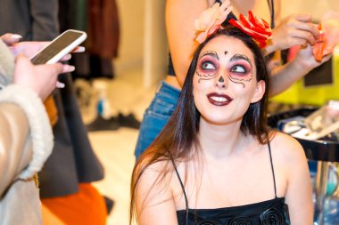 Yüzünde kafatası olan bir kadın kameranın önünde oturuyor. Gülümsüyor ve elinde cep telefonu var. Arkasında başka bir kadın var, fotoğraf çekiyor. Cadılar Bayramı partisi hazırlıkları için.