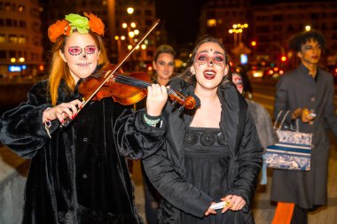 Cadılar Bayramı kostümü giymiş iki kadın keman çalıyor. Biri kemanı tutuyor, diğeri de çelloyu tutuyor. Çevrelerinde, bazıları el çantası tutan başka insanlar da var. Şehirdeki Cadılar Bayramı partisinde.