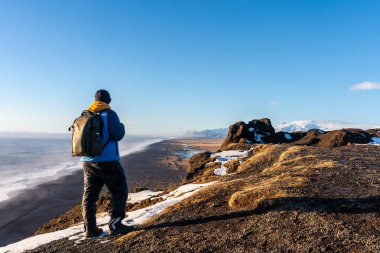 Soğuk kış mevsiminde İzlanda 'da kar yağarken Dyrholaey plajında sırtını dönen bir turist.
