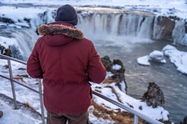 Erkek bir yürüyüşçü kışın gün batımında donmuş Godafoss şelalesine bakıyor, İzlanda.