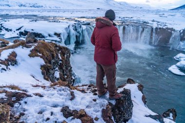 Erkek bir yürüyüşçü kışın gün batımında donmuş Godafoss şelalesine bakıyor, İzlanda.