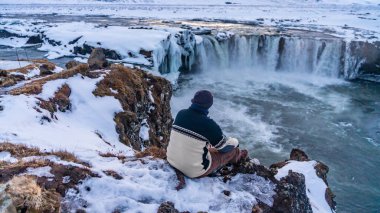 Kışın gün batımında donmuş Godafoss şelalesine bakan bir adam İzlanda 'da oturuyor.