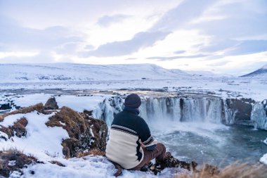 Kışın gün batımında donmuş Godafoss şelalesine bakan bir adam İzlanda 'da oturuyor.
