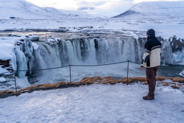 Kışın gün batımında donmuş Godafoss şelalesine bakan bir yürüyüşçü, İzlanda