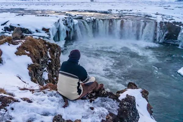 Kışın gün batımında donmuş Godafoss şelalesine bakan bir yürüyüşçü İzlanda 'da oturuyor.