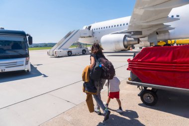 Bir kadın ve bir çocuk beyaz bir uçaktan bavullarıyla uzaklaşıyorlar. Kadın bir sırt çantası taşıyor ve çocuk kırmızı bir gömlek giyiyor.