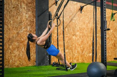 Asyalı güçlü bir kadının modern bir spor salonunda bisepslerini spor yüzükleri kullanarak kullanışının geniş kenarlı görüntüsü.