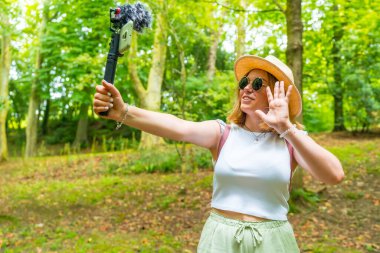 Beyaz tenli bir kadının yatay fotoğrafı. Genç bir kadın. Ormanda yürürken video bloğu kaydediyor.