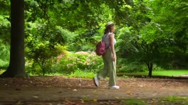 Pembe sırt çantalı bir kadın parkta yürüyor. Park yemyeşil ve yemyeşil, ağaçlar ve otlarla dolu. Kadın hasır şapka ve beyaz ayakkabılar giyiyor. Sahne huzurlu ve rahatlatıcı.