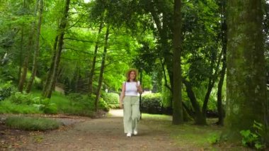 Bir kadın elinde sopayla ve şapkasıyla parkta yürüyor. Park ağaçlar ve otlarla dolu. Huzurlu ve huzurlu bir atmosfer yaratıyor.
