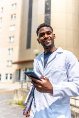 Hastanenin önünde telefonu kullanan neşeli, genç bir Afrikalı doktorun dikey fotoğrafı.
