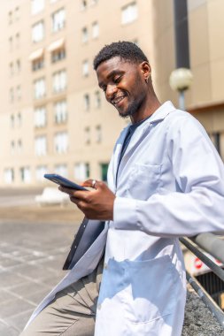 Hastanenin dışında iş molası sırasında telefonla konuşan neşeli, genç bir Afrikalı doktorun dikey fotoğrafı.