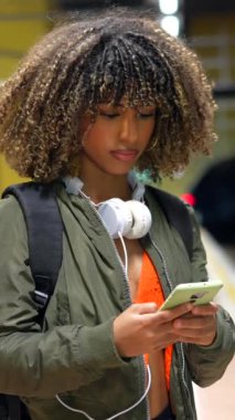 Kıvırcık saçlı ve esmer tenli güzel kadın metro istasyonunu beklerken telefon kullanıyor.