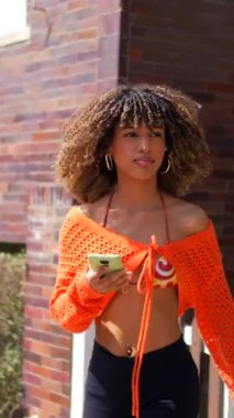 Şehirde telefon kullanırken kıvırcık saçlı Latin bir kız öğrencinin yan görüntüsü.