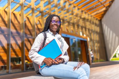 Üniversitenin dışında oturan ve güneşli bir günde kameraya gülümseyen Afrikalı bir kız öğrenci.