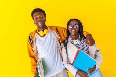 Kameraya sarılıp gülümseyen iki üniversiteli erkek ve kadın Afrikalı meslektaşın sarı arka planlı stüdyo portresi.