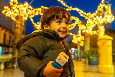 Genç bir çocuk elinde oyuncak bir arabayla bir Noel ağacının önünde duruyor. Sahne neşeli ve neşeli, ağaçtaki ışıklar ve çocuğun gülümsemesiyle