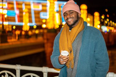 Afrikalı bir adam gecenin karanlığında şehrin Noel ışıklarının yanında kameraya gülümsüyor ve elinde kahve fincanı tutuyor.