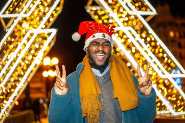 Şehirde Noel 'de gece vakti Noel Baba şapkası takan eğlenceli, havalı bir Afrikalı gencin portresi.