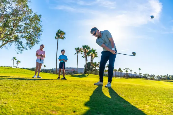 stock image Full length photo of a male caucasian adult golf coach swinging golf next to two young students in a course