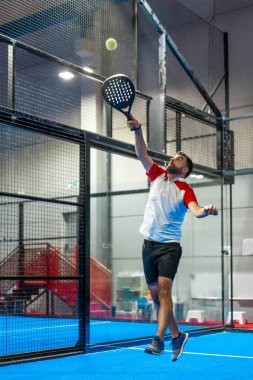 Yetişkin bir adamın topa ulaşmak için zıpladığı ve kapalı mavi bir kortta tenis oynadığı dikey fotoğraf.