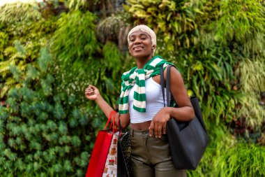 African woman looking at camera while carrying shopping bags next to a garden clipart