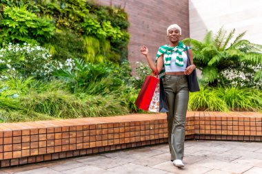 Vertical full length photo of a cool african woman walking carrying shopping bags outside a mall clipart