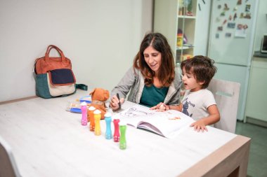 Bir kadın ve bir çocuk önlerinde bir kitapla bir masada oturuyorlar. Kadın çocuğa çizimlerinde yardım ediyor.