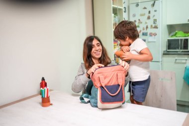 Bir kadın küçük bir çocuğun sırt çantasını açmasına yardım ediyor. Sahne, arka planda mikrodalga fırın ve buzdolabı olan bir mutfakta geçiyor.