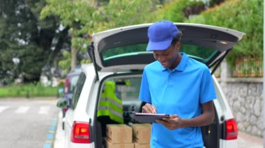 Afrikalı kurye, dijital tablet kullanıyor. Paketleri teslim etmek için minibüsün yanında duruyor.
