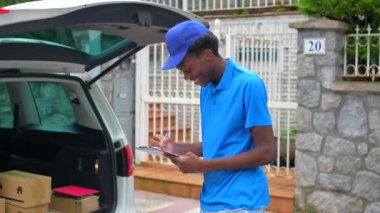 Mavi üniformalı ve şapkalı Afrikalı teslimatçı sokakta bir minibüsün yanında envanter çıkarıyor.