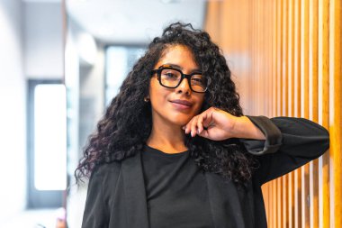 Kendine güvenen, neşeli, uzun kıvırcık saçlı ve modern bir finans binasının içinde kameraya gülümseyen zarif elbiseli bir Latin iş kadınının portresi.