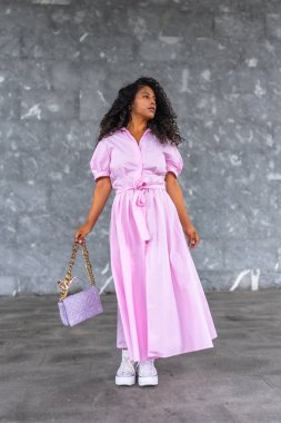 Pembe elbise ve el çantasıyla poz veren şık Latin bir kadının gri şehir duvarına karşı dikey yakın plan portresi