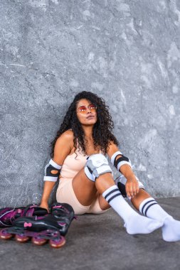 Rahatlamış, sportif, kenar mahalle duvarına yaslanmış patenleriyle oturan Latin bir kadının fotokopi alanı olan dikey portresi.