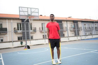 Afrikalı bir adam açık hava mavi basketbol sahasında elastik bant kullanarak ısınıyor.