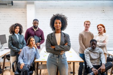 Confident african woman standing with arms crossed next to colleagues in a coworking clipart