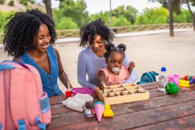 Afrikalı büyükanne, anne ve kız parkta bir piknik masasında oyuncaklarla oynuyorlar.