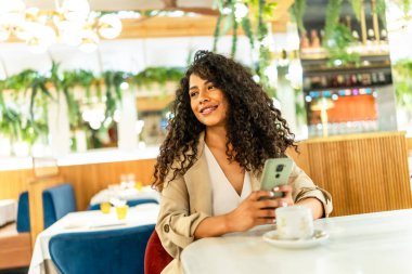 Kafeteryada kahve içerken cep telefonu kullanan dikkati dağılmış Latin kadın.