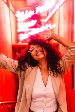 Young and happy latin beauty woman with curly hair dancing near red neon sign clipart