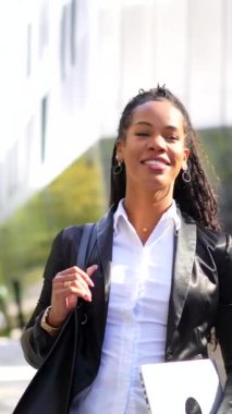 Latin bir iş kadınının şehirde dizüstü bilgisayarı ve el çantasıyla dolaşmasının kopyası olan dikey video.