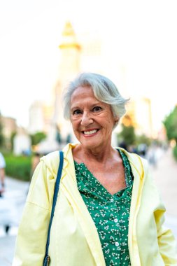 Şehirde kameraya gülümseyen yaşlı ve modern bir kadının dikey yakın plan portresi.