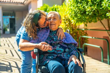 Güneşli bir günde yaşlı kadın bakıcı, yaşlılar bahçesinde tekerlekli sandalyedeki yaşlı kadını öpüyor.