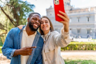 Şehir manzaralı bir parkta selfie çekip gülümseyen çok ırklı mutlu bir çift.