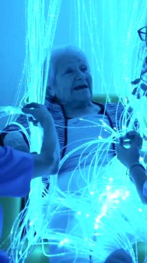 Üst düzey bir kadın, modern bir yaşlılıkta iki bakıcının yanında, duyusal odada ışıklarla oynamaktan zevk alıyor.